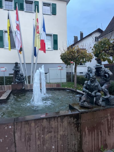 The image depicts a multi-figure bronze fountain situated in Oppenau, Germany. The fountain basin, dark-toned and appearing to be concrete or weathered stone, exhibits vertical dark streaks indicative of water runoff and mineral deposits. Multiple water jets actively propel streams upwards from the basin.

On the left side of the fountain, two bronze statues are positioned: an adult male figure stands, holding a long, slender object (possibly a tool or staff), while a smaller figure (child or youth) is seated at his feet, looking towards the standing figure. On the right side of the fountain, three additional bronze statues are grouped. A male figure is seated, wearing an apron, actively engaged in a task with a tool in front of him. Behind him, a standing male figure wears a hat and holds a cylindrical container, while a partially obscured female figure, wearing a head covering, stands between the two male figures.

In the background, a light-colored, multi-story building features several windows with dark green louvered shutters. To the right of this building, another white-facaded structure with a dark, sloped roof and two dark chimneys is visible.

Five flags are displayed on individual metal poles positioned in front of the primary building. From left to right, these include a yellow and black flag, a white flag with a red crest, a partially visible German flag (black, red, gold), a yellow flag with red text, and the French national flag (blue, white, red).

Two informational placards on metal stands are present near the fountain; one on the left displays a portrait. A gray utility box is located behind the fountain, to the right of the flags. The surrounding environment includes small, deciduous trees with sparse, yellow-brown autumnal foliage, and a grassy area. The sky is uniformly overcast.