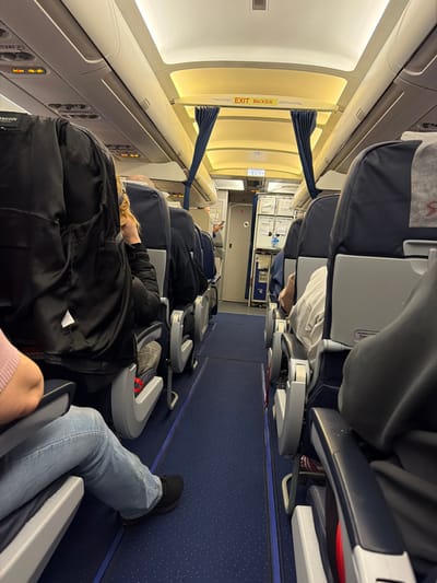 This image depicts the interior cabin of a commercial aircraft, presumably at Емельяново (аэропорт), Russia. The perspective is from a passenger seat, looking forward down the main aisle towards the front of the aircraft.
**Environment:**
The aircraft cabin features rows of blue and gray passenger seats arranged in a typical 3-3 or 2-2 configuration (specific number per row not fully visible). A narrow blue carpeted aisle runs down the center. Overhead bins are closed and illuminated by warm white LED lighting, which also illuminates the ceiling. An "EXIT ВЫХОД" sign, displayed in both English and Russian, is centrally positioned above the aisle in the forward section of the cabin. Dark blue curtains are pulled back on either side of this exit sign. Further forward, a galley or cabin crew station is visible, containing various panels and equipment.
**People:**
Multiple passengers are seated throughout the cabin. In the immediate foreground on the left, a person is seated, wearing light blue jeans and black athletic shoes. Their right arm, covered by a light pink long-sleeved garment, rests on the armrest. The head and torso of this individual are out of frame. The seat directly in front of this person is occupied by an individual in a black jacket, their head partially visible with blonde hair. Further down the left aisle, several other passengers are visible, primarily from the back of their heads and shoulders, occupying seats. On the right side of the aisle, a passenger is seated in the foreground, primarily visible from their right arm and side, wearing a light gray long-sleeved shirt and dark trousers. Other passengers are visible further down the right aisle, similarly seated. No clear active interactions between passengers are discernable.
**Objects:**
Identifiable objects include passenger seats with blue fabric and gray plastic frames, overhead bins, illuminated ceiling panels, the "EXIT ВЫХОД" sign, blue curtains, and the blue speckled carpet. The seatbacks have literature pockets and a small flip-down tray table (not in use). A logo, possibly "S7 Airlines," is faintly visible on the headrest cover of one seat on the right. A small, dark object (possibly a cell phone) is held by a person in the forward galley area on the left.
