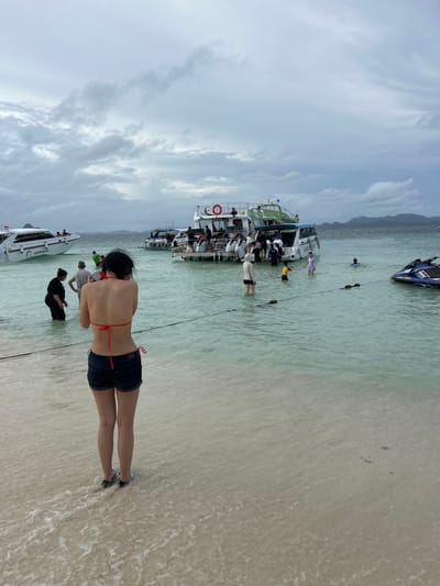 The image depicts a crowded beach scene in Tambon Phru Nai, Thailand. The water is clear and shallow, with several boats anchored offshore, including speedboats and a larger passenger boat. Many people are in the water, some wading and others swimming. On the beach, a woman in a bikini and denim shorts is facing away from the camera, looking out at the water. In the background, a jet ski is visible near the shore. The sky is overcast with scattered clouds, suggesting a moderate weather day. The overall impression is one of a popular tourist destination with active water-based recreation.