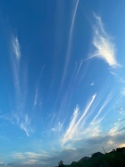 The image depicts a clear blue sky with wispy, white cirrus clouds stretching across it. The clouds are elongated and appear to be blown by the wind, creating a dynamic and streaky pattern. The lower portion of the image shows a silhouetted landscape with dark green grass and the dark shapes of hills or mountains. The overall scene suggests an outdoor environment, possibly a rural or semi-rural setting, under a bright sky. There are no people or specific man-made structures visible in the image that would indicate a precise location within Manhush, Ukraine, or any other city.