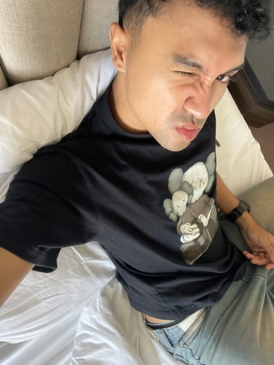 An overhead photograph taken in Kayuambon, Indonesia, shows a young adult male lying on his back on a bed, looking directly into the camera. He has a medium complexion, short dark curly hair, and is winking with his left eye, with his nose crinkled and lips slightly pursed. He is wearing a black short-sleeved t-shirt with a graphic print of three KAWS Companion figures on the chest: two smaller grey figures and one larger brown figure. A black digital watch is visible on his left wrist. Below the t-shirt, light blue denim jeans are visible, with the light blue waistband of an undergarment peeking out above them. The bed is covered with white sheets and a white pillow supports his head. A light-colored, possibly beige or cream, upholstered headboard or cushion is behind him to the left. The scene is indoors, likely a bedroom or hotel room.