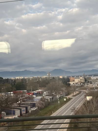 An elevated, wide-angle view captures an industrial and commercial landscape within Vilobí d'Onyar, Spain. The upper portion of the frame is dominated by an overcast sky with heavy cloud cover. Two prominent, bright, rounded rectangular reflections are visible in the upper-mid section, indicating the photograph was taken through a window, possibly from a moving vehicle. Faint contrails are also discernable within the cloud layer.

In the immediate foreground, a dark metal railing runs horizontally across the bottom. The midground on the left features a large, unpaved lot filled with numerous parked semi-trailer trucks and their trailers, some marked with "EDY" branding. Deciduous trees, mostly bare, are scattered throughout the lot and along its perimeter, with one tree exhibiting early pink blossoms. Low-rise, grey industrial or commercial buildings are situated behind the truck parking area.

To the right, a two-lane paved road with white lane markings curves through the scene, appearing to merge or intersect with other routes. Several vehicles, including cars and trucks, are sparsely distributed on or near the road. A multi-story, light brown building with the word "HOTEL" clearly visible on its side stands prominently beside the road, accompanied by a small parking area. A billboard is partially visible on the far right.

In the background, a sprawling complex of buildings extends towards the horizon, culminating in a distinct airport control tower located in the central background. A range of blue-grey mountains forms the distant horizon line. The scene is largely static, with no discernible individuals.