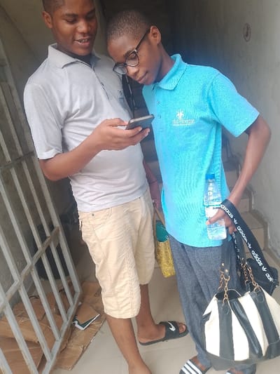 Two young males are depicted standing indoors or in a covered outdoor area in Mgbuoshimini, Nigeria. The male on the left, wearing a light grey polo shirt and beige shorts, holds a dark smartphone horizontally in his right hand, looking down at the screen with his mouth slightly ajar. He wears dark slides on his feet. The male on the right, wearing black-rimmed glasses, a light blue polo shirt with a "CLUB ROYAL UMUAHIA" logo, and dark grey sweatpants, leans towards the first male, also looking at the smartphone. He holds a clear "Jozza" brand plastic water bottle in his left hand and a black and white striped shoulder bag with an "adidas" branded strap in his right hand. A gold-colored fabric bag is also visible on his left side. He wears white and black striped slides. The background features a light-colored wall, a light-colored metal gate or railing to the left, and a floor with visible cardboard boxes and a dark book near the gate. Steps are partially visible on the right.
