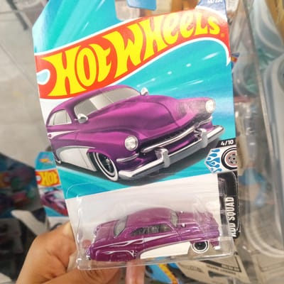 A person's hand holds a blister pack containing a purple Hot Wheels toy car. The car is a classic model with white accents and whitewall tires. The background shows a blurry retail environment, suggesting the items are on display in a store. The Hot Wheels logo is prominently featured on the packaging, and there are other toy cars and retail displays visible in the background. The scene does not provide any information about the specific location within El Limon, Venezuela, as it is a generic retail setting.