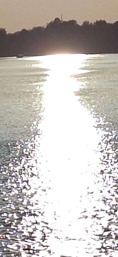 The image captures a sunlit water surface, likely a river or bay, with a distinct bright reflection of the sun creating a shimmering path. In the distance, a silhouetted treeline forms the horizon, with a slender, pointed structure, possibly a spire or tower, barely visible against the sky. The water exhibits a rippling texture, with small whitecaps or light reflections scattered across it, particularly in the foreground. The overall impression is one of a bright, possibly early morning or late afternoon scene, with the sun's glare dominating the visual field. The location is specified as Pärnu linn, Estonia.