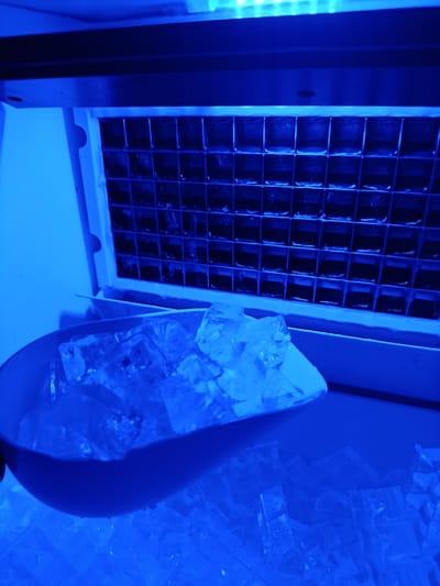 A close-up, overhead shot of an ice dispenser and scoop filled with ice cubes, illuminated by a bright blue light. The ice dispenser grid is visible in the background. The scene is set in Pishchane, Ukraine. There are no people or animals present in the image. The focus is on the ice and the dispenser mechanism.