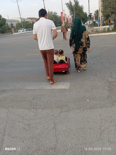 In Bekabad, Uzbekistan, on June 16, 2026, at 19:20, a man in a white polo shirt and brown pants walks across a pedestrian crossing, pushing a red toy car with two children inside. A woman in a camouflage-patterned outfit and a dark green hijab walks behind him. In the background, traffic lights show green, indicating it is safe for vehicles to proceed. The scene unfolds on an asphalt road with buildings and trees lining the sides.