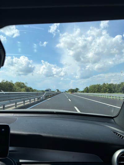 The image captures a view from inside a vehicle driving on a highway. The perspective is from the passenger side, looking forward and slightly upwards. The scene is dominated by a bright blue sky filled with fluffy white clouds, suggesting pleasant weather. The highway itself is paved and appears to be a multi-lane road, indicated by the white lane markings. On the right side of the road, a metal guardrail runs parallel to the highway, separating it from a dense line of green trees. The trees form a natural barrier along the edge of the road, characteristic of a rural or semi-rural setting.
Inside the vehicle, the dashboard is partially visible on the right side of the frame, consisting of dark, textured plastic. A portion of the car's window is also visible, showing some smudges and reflections. In the distance, along the highway, other vehicles can be seen, and there are some overhead structures that might be gantries for traffic signs or toll booths. The overall impression is of a journey on a sunny day, likely in the daytime, given the brightness of the sky.
There is no visible text in the image. The provided location context, Chiasottis / Cjasotis, Italy, suggests the highway is located in this region. The image conveys a sense of movement and travel.