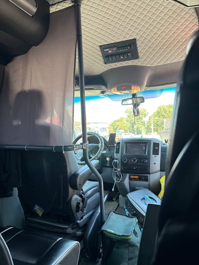 The image shows the interior of a van or truck, viewed from the passenger seat. The driver's seat is visible on the left, upholstered in black and gray material, with a black mesh pocket on the back. A grey curtain is drawn across the view towards the front of the cabin, obscuring the driver's head and torso. Above the curtain, the ceiling is upholstered in a diamond-quilted pattern. A panel with several buttons and a digital display is mounted on the ceiling. The dashboard is visible, featuring a touchscreen infotainment system, air vents, and climate controls. A phone mount with a phone is attached to the dashboard. On the passenger side, a black pet carrier with light blue accents and a green cooler bag are on the floor. Outside the vehicle, through the windshield, trees and buildings are visible under a bright sky. The scene is a static interior view of a vehicle, likely parked.
