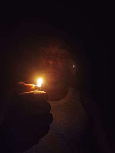 An image shows a person holding a lit lighter in front of their face. The background is dark, suggesting the photo was taken at night or in a dimly lit environment. The person is wearing a white tank top. A green reflection is visible on the person's face. The lighting from the lighter illuminates the hand, part of the face, and the tank top. The overall impression is a close-up shot focused on the action of lighting the lighter. The location of Orodo, Nigeria is not discernible from the image.