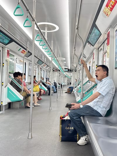 The image captures the interior of a modern subway car in Tiaodeng, Chongqing, China. The carriage is brightly lit with white walls, a grey floor, and features light teal accents on the seating and hanging grab handles. Silver poles and rails run throughout the cabin, providing support for standing passengers. Digital screens are visible above the windows, and warning signs are affixed to the walls.
In the foreground, a man with short, dark hair, wearing a light blue striped shirt, blue jeans, and white Nike sneakers, is seated on a light grey bench. He holds onto a silver grab rail with his left hand, his mouth wide open in what appears to be a large yawn or shout, creating a candid and dramatic expression. He wears a silver watch on his left wrist and holds a gold-colored smartphone in his right hand. Next to his feet on the grey floor is a dark blue shopping bag with gold Chinese characters that read "青花汾酒" (Qinghua Fenjiu), suggesting a purchase of liquor.
Further down the subway car, several other passengers are visible, mostly seated and either looking at their phones or resting. The overall scene depicts a routine journey on public transport.
Visible text includes safety warnings such as "▲一当心夹手 Warning hands pinching" and "Fire Extinguisher Operation" alongside Chinese characters "灭火器操作说". Other signage welcomes passengers with "十八号线欢迎您" (Welcome to Line 18) and identifies the line as "重庆轨道十八号" (Chongqing Rail Transit Line 18).