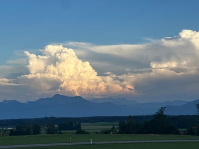 The image captures a wide-angle view of a rural landscape under a dramatic sky, likely taken from the vicinity of Höslwang, Germany. Dominating the upper half of the frame are large, volumetric cumulus clouds, illuminated by the warm tones of setting sunlight, with a clear blue sky visible above. These clouds are situated in front of a layered mountain range, rendered in deep blue and purple hues due to atmospheric perspective.
Below the mountains, a dark band of trees forms a dense forest line, separating the mountains from a foreground of rolling green fields. The fields are interspersed with scattered trees and what appear to be farm buildings. A road with white lines runs horizontally across the lower part of the image. In the middle ground of the fields, two cows, one brown and white and the other darker, are visible. The scene is devoid of any human presence or discernible action. The overall impression is one of serene natural beauty at dusk.