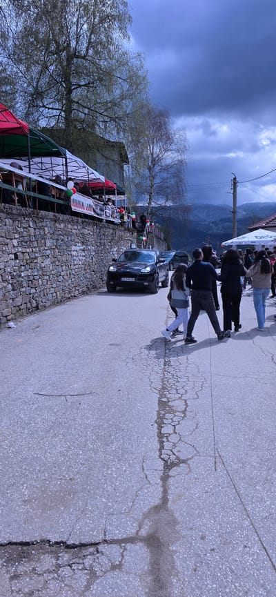 The image shows a street scene in Breze, Bulgaria. A group of people are walking on a paved road, seemingly participating in an event or parade. A black car is parked on the side of the road, next to a stone wall. Behind the wall, there is a building decorated with flags and balloons. The sky is cloudy and a mountain range is visible in the background.