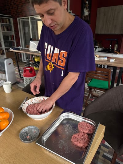 A man wearing a Phoenix Suns T-shirt is making hamburger patties. He is mixing ground beef in a white bowl. To his right is a metal tray holding two hamburger patties. Next to the bowl is a hamburger press and a bowl of oranges. The location is Ptichnoye, Russia.