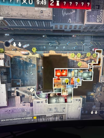 The image displays an aerial, top-down perspective of an urban environment within the city of Vellmar, Germany, as presented on a digital screen. The scene focuses on a central building complex surrounded by streets and other structures.
**Environment:**
A wide, multi-lane street stretches across the upper portion of the view, populated by approximately two dozen vehicles, including sedans, trucks, and vans. Many of these vehicles appear damaged, overturned, or irregularly positioned across the roadway. Two translucent white human-silhouette icons are visible on this main street near a black circular void. Sidewalks with trees and lampposts border the street. To the left, a narrower street runs alongside a parking lot filled with additional vehicles. Numerous multi-story buildings with flat roofs and visible window patterns line the streets, forming a dense urban block.
**Central Complex:**
The prominent central building complex is rendered with an "x-ray" effect, exposing its interior layout across multiple floors. The internal rooms are depicted in various colors, including red, white, and light blue, containing representations of furniture such as tables and beds. Two distinct yellow markers, labeled "A" and "B," are prominently positioned within separate rooms on an upper floor of this complex.
**Digital Overlays and Objects:**
At the top of the screen, digital overlays indicate a scoreboard of "0" versus "2," a timer displaying "9:49," and the text "MATCHBALL." Red question marks are visible adjacent to a red chess rook icon. At the bottom left, a yellow briefcase icon is accompanied by the German text "DEN ENTSCHÄRFER AUFHEBEN" (PICK UP THE DEFUSER). Faint branding "AOC" is visible at the very bottom edge of the screen, along with a partial Windows activation watermark ("Windows aktiv...").