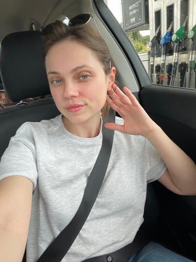 A young woman is seated in a car, wearing a light grey t-shirt and a seatbelt. Her left hand is raised near her ear. Sunglasses are perched on her head. A gas station with multiple pumps is visible through the car window. The setting appears to be a gas station in Perugia, Italy.
