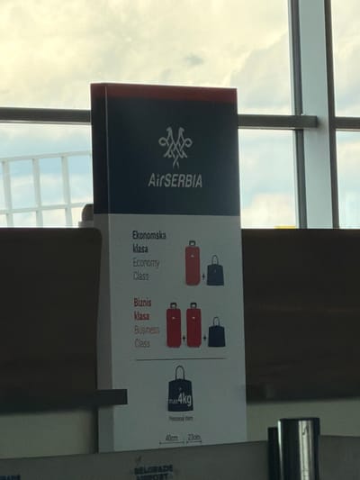 This media file captures an informational sign for Air Serbia within what appears to be an airport terminal. The primary subject is a large, upright sign featuring the Air Serbia logo and text on a dark blue or black background, with the bulk of the information presented on a white surface. No people are visible in the image.
The setting is an indoor space with large windows in the background, revealing an overcast sky with clouds. This suggests it is daytime. The sign itself is the main object, displaying baggage allowance rules. It shows visual representations of luggage: one red suitcase and one black personal item for "Ekonomska klasa / Economy Class," and two red suitcases and one black personal item for "Biznis klasa / Business Class." A notable detail is the maximum weight specified for the personal item as "max 4kg," along with "Personal item" and dimensions "40cm 23cm."
The visible text includes "Air SERBIA," "Ekonomska klasa," "Economy Class," "Biznis klasa," "Business Class," "max 4kg," "Personal item," "40cm," and "23cm." Partially visible at the bottom right is "BELGRADE AIRPORT," confirming the location as Belgrade Nikola Tesla Airport in Surcin Urban Municipality, Serbia. The image itself is rotated 90 degrees clockwise, making the sign appear horizontal in the frame. No specific activity or event is taking place; the image simply documents the static informational display.