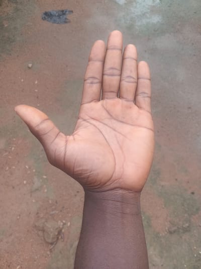 A close-up shot displays the palm and part of the forearm of a dark-skinned individual. The hand is held palm-up, fingers slightly spread, revealing the natural lines and creases of the palm. The skin tone is a deep brown, with some areas appearing lighter due to the lighting and natural variations. The forearm, visible on the right side of the frame, transitions to a darker hue.

The background is a textured, earthy surface, appearing to be a mix of dirt and concrete or packed earth. The colors in the background are predominantly muted browns and grays, with some lighter, mottled patches. To the far left, a dark, indistinct smudge or shadow is present on the ground, its form unclear. The lighting suggests it could be daytime, with diffused light casting soft shadows. There is no visible text in the image. The overall impression is of a candid, everyday moment captured in an outdoor setting, likely in Gbaza, Nigeria, given the context. The image is devoid of any specific activity or event beyond the static pose of the hand.