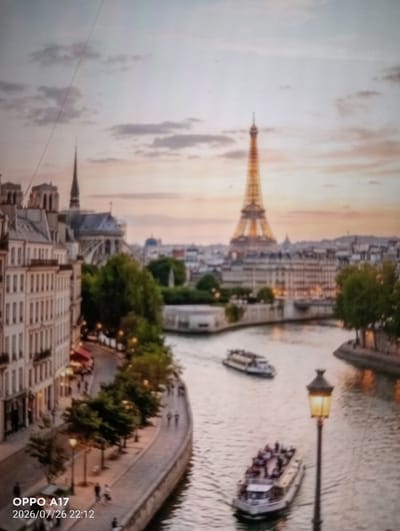 The image depicts a scenic view of Paris, France, specifically the Seine River with the Eiffel Tower visible in the background. The photograph captures a city vista at dusk or dawn, with warm light illuminating the iconic landmark and the surrounding buildings. Several tour boats are navigating the river, carrying passengers. Along the riverbank, there are trees and lampposts, suggesting a promenade or park area. The architecture of the buildings lining the river is typical of Parisian style. The timestamp in the lower left corner indicates "OPPO A17 2026/07/26 22:12," which is inconsistent with the visual content of Paris. The question specifying the city of Құмөзек, Kazakhstan, is therefore incongruous with the image provided.