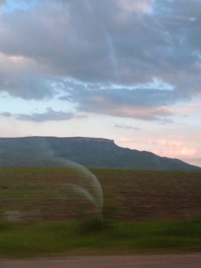 The image depicts a rural landscape with a flat-topped mountain range in the distance under a sky filled with clouds. The foreground consists of a field with green grass and brown soil, and a water sprinkler arm is visible creating a curved arc of mist over the field. The lighting suggests it is either early morning or late evening. There are no people or man-made structures immediately apparent in the scene, though the presence of a sprinkler suggests agricultural activity. This scene is consistent with the rural environment surrounding Storozhevaya, Russia, where such landscapes are common.
