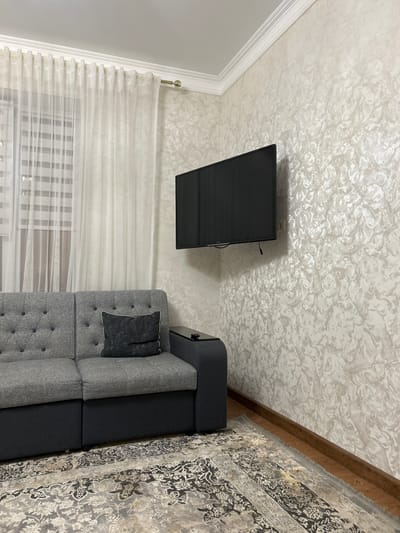 A medium shot captures a living room scene with a modern aesthetic. The primary focus is a gray sectional sofa with tufted cushions and a single dark gray decorative pillow. To the right of the sofa, a black side table with a remote control is visible. Mounted on the wall above the side table is a flat-screen television. The wall itself is textured with a light, abstract pattern in shades of beige and off-white.
To the left of the sofa, sheer white curtains are drawn, partially obscuring horizontal blinds on a window. The floor is covered by a large area rug with an ornate, traditional pattern in muted tones of gray, beige, and black. The room is illuminated by soft, ambient light, suggesting an indoor setting. The environment appears to be a residential living space, possibly in the city of Nizhneye Kazanishche, Russia, given the provided location context. There are no people visible in the image.