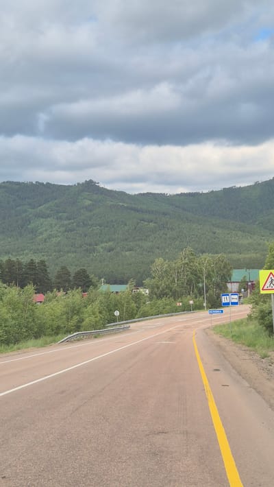 The image depicts a view of a road curving to the right, leading towards the village of Elovka, Russia. The road has a light brown asphalt surface with a white line marking the left edge and a bright yellow line marking the right lane. On the right side of the road, there are several signs: a yellow diamond-shaped sign with a pedestrian silhouette, a blue rectangular sign with a bed icon indicating accommodation, and another blue rectangular sign with a bed icon and what appears to be a bus icon. A smaller blue sign with Cyrillic text, "Еловка," is also visible. The surrounding environment features lush green trees and dense forest on a hillside. In the distance, a large, heavily forested mountain range dominates the landscape under a cloudy sky. Several buildings with red and green roofs are visible amidst the trees in the mid-ground. A metal guardrail runs along the left side of the road. The scene suggests a rural setting with a paved road passing through a forested area and approaching a village.