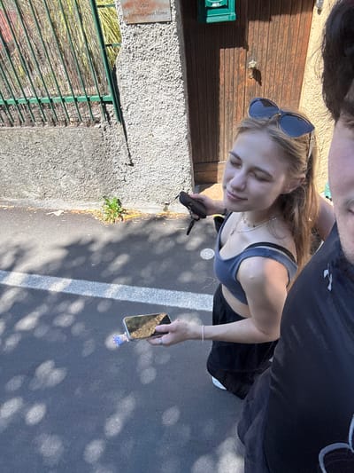 An outdoor, eye-level, medium shot shows two individuals near a textured wall and a wooden door. The individual on the left, a young woman, is facing right with her eyes closed and a slight smile, wearing sunglasses on her head, a blue tank top, and a black skirt. She holds a black cell phone in her left hand. The individual on the right, partially visible and appearing to be male, is facing forward, wearing a black shirt. A white stripe is visible on the asphalt ground in front of them. A green mailbox is mounted on the wooden door. A sign above the door reads "VSH Varenna Sweet Home." The scene is illuminated by sunlight, creating shadows on the ground.