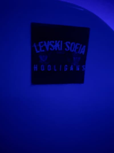 A black rectangular sticker with blue text is adhered to a dark blue surface, possibly a wall or a curved object. The sticker contains two lines of text. The top line reads "LEVSKI SOFIA" in a stylized font. Below this, a crest or logo is visible, followed by the word "HOOLIGANS" in a similar blue font to the top line. The sticker appears to be illuminated by a blue light source, which gives the entire scene a predominantly blue hue. The background is a deep, rich blue. The image is a close-up, with the sticker being the central focus. No people or other objects are visible. The location in Paralimni, Cyprus is not discernible from the image itself.