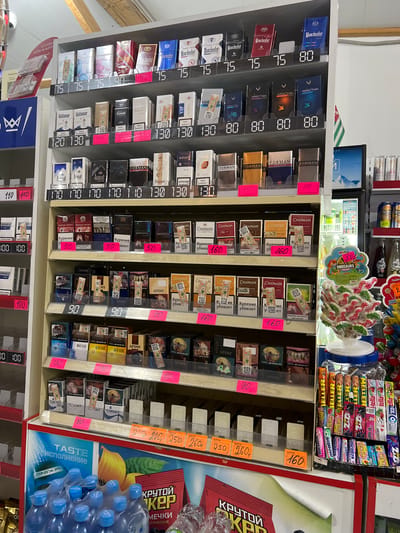 This image depicts a retail display of cigarettes and confectioneries inside a store, likely a convenience store or kiosk, located in Pitsunda, Abkhazia. The display shelves are organized with various brands of cigarettes, each with a digital price tag. Brands visible include Manchester, Arizona, Forman, Elegant, APsnY, Esse, and Chapman. Among the confectioneries are Meller candies and Orbit gum.
In the foreground, a multi-pack of bottled water is visible. A refrigerated display case, possibly for beverages or chilled items, can be seen in the background. A small flag, appearing to be the flag of Abkhazia, is also present in the background. The lighting suggests an indoor environment, with fluorescent lights illuminating the products. There are no people visible in the image, and the focus is solely on the merchandise and the store's interior. The overall scene conveys a commercial setting where goods are presented for sale.