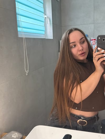 A young woman with long, brown hair is taking a selfie in a bathroom. She is wearing a brown crop top and dark jeans with a belt. The bathroom has grey tiled walls and a window with blue blinds. A pearl necklace with a small charm hangs on the wall to the left of the woman. A white sink is visible in the foreground. There are also some toiletries on a shelf behind her. The image is taken from the chest up, with the woman's face and upper body being the main focus. There is no indication of the city of Kamyennoye, Russia in the image.