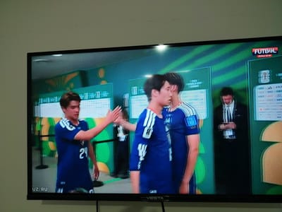 The image depicts a television screen displaying a sporting event. On the screen, a young man in a blue jersey with the number 24 is high-fiving another person. Both are wearing blue jerseys with white accents. In the background, other individuals are visible, one in a suit and tie, and another wearing a dark outfit. The backdrop appears to be a green and yellow patterned wall, possibly representing a sports venue or a broadcast studio. A logo in the upper right corner reads "FUTBOL" in red. At the bottom left of the screen, "UZ RU" is visible, suggesting a broadcast language or region setting. The screen itself is mounted on a black frame, and the image is captured from an angle, with a slightly out-of-focus view of a white wall in the foreground. The overall scene is a captured moment from a televised sports broadcast, likely a football match. The presence of "UZ RU" could indicate that this broadcast is being shown or is relevant to Uzbekistan, which has Termez as one of its cities.