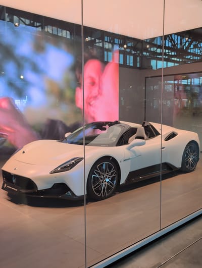 The image shows a white Maserati MC20 Cielo sports car on display inside a modern showroom, likely at the Motorworld Region Stuttgart in Böblingen, Germany. The car is positioned centrally, facing left, with its convertible roof down, revealing the interior. The showroom features a sleek, minimalist design with large glass windows and a grey tiled floor. Behind the car, a large digital screen displays a blurred, abstract image of human figures and colorful lights. Reflections of the showroom's ceiling and industrial-style architecture, including exposed beams and lighting fixtures, are visible in the glass and on the car's surface. The car has black accents on the wheels, side skirts, and front splitter. A "MASERATI APPROVED" plaque is visible on the lower front of the car. The overall scene suggests a high-end automotive display or dealership.