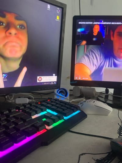 The image shows a desk setup with two computer monitors, a keyboard, and a mouse. The top monitor displays a video with a man's face in the foreground and another man's face in a smaller window. Text on the screen reads "ЗУБАРЕВ СМОТРИТ «АМЕР ПИРОГ ВСЕ В СБОРЕ»". The bottom monitor displays a desktop with icons, including one for "Rocket League". It also shows a notification at the top right with the date "30.06.2020". The keyboard is a gaming keyboard with RGB lighting. A white gaming mouse is also visible. The overall scene appears to be a home office or gaming setup. The lighting suggests it could be daytime, but there are no direct indications of weather. The presence of the "Rocket League" icon and the gaming keyboard suggests the activity taking place is likely gaming or related computer use. The image is oriented vertically.