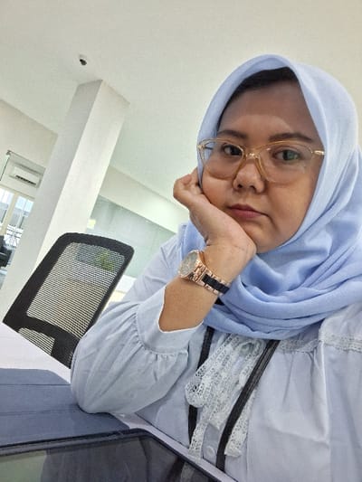 A person wearing a light blue hijab and gold-framed glasses rests their head on their hand, wrist adorned with a two-tone watch. They wear a light blue top with white lace details. A black mesh office chair is visible in the background, as well as a white wall. The angle suggests the picture was taken at a tilted perspective.