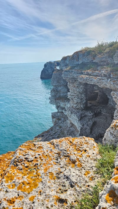 The image shows a coastal landscape near Kamen Bryag, Bulgaria. In the foreground, a rugged, light-colored cliff face is covered with patches of orange lichen and small green plants. The cliff edge extends over the turquoise sea. Behind this, there's a deeper, textured cliff formation with a cave opening. In the distance, a sea stack with a natural arch is visible. The sky is pale blue with faint cloud trails. The perspective is from a high vantage point.