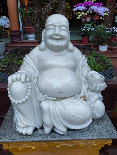 A white stone statue of a seated, laughing Buddha (Hotei) is positioned prominently outdoors, resting on a two-tiered pedestal with a dark grey upper surface and a gold-colored lower section. The statue features a bald figure with a pronounced belly, a wide smile, and closed eyes. Its left hand holds a string of white prayer beads, while its right hand holds a white object resembling a gold ingot. Minor surface discoloration and dirt are visible on the statue.
Behind the statue, a narrow, dark planter box contains dark soil and sparse green sprouts. Further back, a paved area leads to a building entrance with reddish-brown pillars and steps. A large tree wrapped with white string lights stands to the left of the building's center. Numerous potted plants are arranged on the steps and surrounding area, including a prominent display of purple and white orchids in bloom, alongside other green foliage plants. Small red decorative elements are interspersed among the flowers. In the upper left background, partially obscured by a pillar, a person wearing a light-colored hooded garment is seated on a rattan chair. The scene depicts an outdoor decorative setting, likely a temple courtyard or garden, in Thành phố Quảng Ngãi, Vietnam.