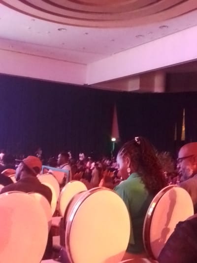 Captured in what appears to be an auditorium or large hall in Satellite Town, Nigeria, a considerable crowd of people are seated, facing a stage or performance area that is largely obscured by the audience and a dark backdrop. Seating appears to consist of round-backed chairs with light-colored cushions and metallic frames. The lighting in the room casts a somewhat pink hue overall, and the ceiling features recessed lighting and a decorative, concentric design. A few vertical objects, possibly banners or light fixtures, are visible on the stage. In the foreground, we see the backs of heads and shoulders of seated attendees, including one woman with intricately braided hair wearing a green shirt and an older man with glasses. There is no discernible action or interaction taking place among the visible attendees.