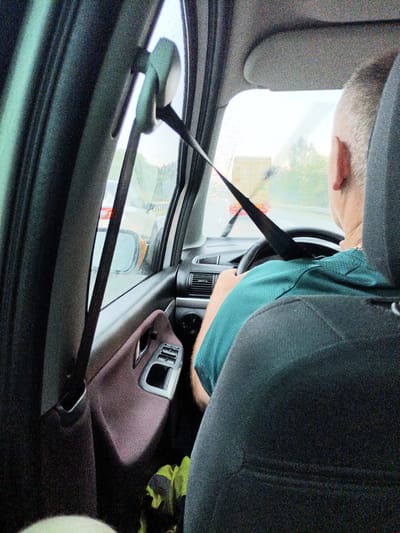 The image shows the interior of a car, viewed from the back seat, as if the photographer is a passenger. The primary subject is the driver, a man with short, light-colored hair, seen from the back and right side. He is wearing a teal polo shirt and is actively driving, with his hands on the steering wheel. His right ear is visible.
The setting is a car interior with a focus on the driver's side. Visible car parts include the steering wheel, dashboard elements like air vents, a car door panel with window controls and a door handle, and a seatbelt which is diagonally across the frame, obscuring part of the driver. The driver's headrest is also visible in the foreground.
The scene takes place during the day, with overcast or rainy weather, indicated by raindrops on the car windows. The view through the windshield shows other cars on a road, suggesting the car is in traffic or on a highway. One car in front is visible with its taillights illuminated, and a larger vehicle, possibly a truck or van, is further ahead. The overall mood is mundane, likely a commute or travel.
There is no visible text in the image. The location context is Divišov, Czechia, which is a small town in the Central Bohemian Region of the Czech Republic. This information, however, cannot be inferred solely from the visual content of the image.