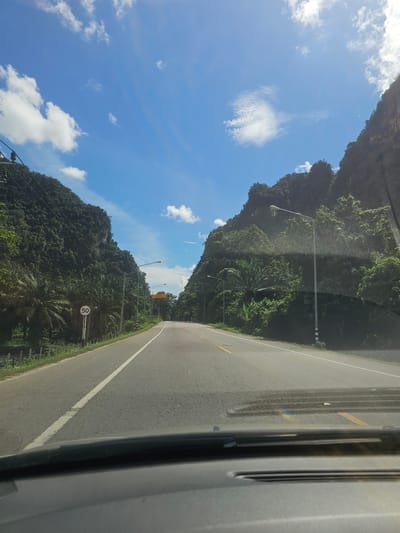 The image shows a view from inside a car driving on a paved road. The road is bordered by steep, lush green mountains on both sides, with a clear blue sky overhead dotted with white clouds. There are utility poles and streetlights lining the road. A speed limit sign indicating "50" is visible on the left side of the road. The scene appears to be set in a natural, mountainous environment. Based on the context provided, the location is the city of Ban Thung Aok, Thailand.