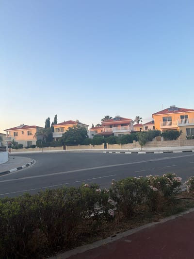 In Pervolia, Cyprus, a street curves to the left, bordered by a sidewalk with black and white painted curbs. On the right side of the street, a row of residential houses with orange-tiled roofs and light-colored walls are visible. Solar panels are installed on the roofs of some houses. Lush green hedges and trees surround the properties. In the foreground, on the right, a patch of flowering bushes with pink and white blossoms grows along the sidewalk. The sky above is a clear, pale blue, indicating daylight. A single person, a woman wearing a light-colored dress, is walking on the sidewalk on the left side of the street, in the distance. The overall scene depicts a quiet residential street on a clear day.