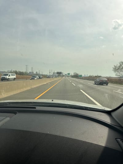 The image presents a first-person view from inside a vehicle, looking out through the windshield onto a multi-lane highway. On the right side of the frame, a dark, textured car dashboard or interior panel is visible, with a shadow cast across a portion of it. The windshield shows several dust particles and smudges.
The highway stretches into the distance, with multiple lanes marked by white dashed lines. A concrete barrier, outlined by a solid yellow line, separates the main lanes from an adjacent lane on the left where a queue of cars indicates moderate to heavy traffic. Other vehicles are also seen traveling in the main lanes.
Along the horizon, trees line the sides of the highway, and faint outlines of distant structures, including power lines or utility poles, are visible. The sky is overcast and hazy, appearing light gray with some subtle cloud formations. The scene depicts a daytime drive on a freeway, likely in or near Ridgefield, USA, during cloudy weather. No specific people, emotions, or legible text are identifiable in the image.