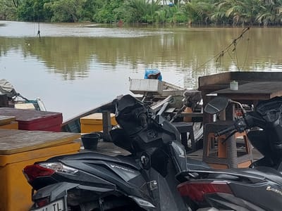 In Parit Baru, Indonesia, a murky brown river flows alongside a dense green jungle. Moored boats are visible on the water, one with a blue tarp covering its contents. On the bank, several yellow and red storage containers are stacked, and two dark-colored motorcycles are parked in the foreground, partially obscuring a wooden table and chairs.