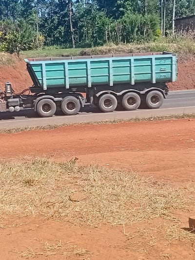 A green truck with six wheels is parked on the side of a road. The road is gray. The area to the right of the truck is dirt-covered, with dried grass and some sparse vegetation. To the left of the truck, there are many trees with green leaves.