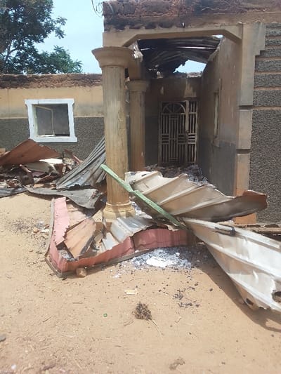 The image shows the exterior of a damaged building in Kwal, Nigeria (latitude: 9.804, longitude: 8.647). The building appears to be a residential home. The front porch is partially collapsed, and the roof is missing in some areas. The front door is made of metal bars and is still in place. The walls are made of brick and concrete. The ground is covered in debris, including broken pieces of roofing material. There are two columns supporting the roof, and one appears to be partially broken. There are no people present in the image.