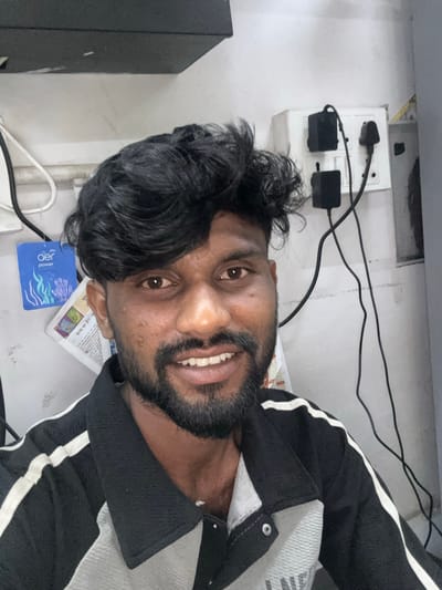 An adult male with dark skin, dark voluminous hair, and a full beard and mustache is depicted in a head-and-shoulders portrait, smiling and looking directly at the camera. He is wearing a black polo shirt with white stripes on the shoulders and a grey upper front section. The indoor background, consistent with a location in Palghar, India, features a light-colored wall. To the subject's right, a white electrical switchboard is visible with multiple switches and two black power adapters plugged in, trailing black cables. To his left, a blue "aer power pocket" air freshener hangs on the wall, and beneath it, several papers with visible Hindi text are affixed. A dark, flat surface is partially visible across the top edge of the frame.