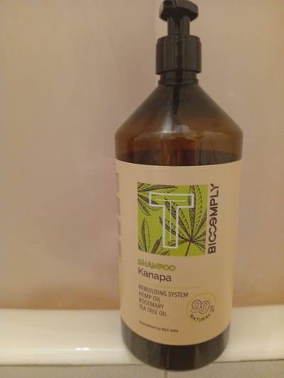 The image shows a bottle of shampoo with a dispenser pump. The bottle is made of amber glass and has a label with text and graphics. The label features a large white letter "T" on a green background with drawings of hemp leaves. The text on the label reads "SHAMPOO Kanapa", "REBUILDING SYSTEM HEMP OIL ROSEMARY TEA TREE OIL", "Guaranteed by BKA Italia", and "BICCOMPLY". There is also a circular emblem with "99% NATURAL" written on it. The bottle is placed on a white surface, possibly a shelf or the edge of a bathtub, against a plain, light-colored wall. The lighting appears to be indoor, and there are no people or other objects prominently visible. The overall scene is a product shot of the shampoo bottle.