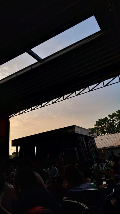 The image shows an outdoor scene in Ogba, Nigeria, likely taken from beneath a partially covered area with a view of the sky. A metal structure with a rectangular opening frames a section of the sky, which transitions from a pale blue to a yellow-orange hue, suggesting either sunrise or sunset. To the right of the metal structure, there is the edge of a building and some foliage. In the foreground and to the right, are indistinct figures of people, some seated at what appears to be a table with bottles and other items, suggesting an informal gathering or dining area. The area is dimly lit, with significant portions in shadow.