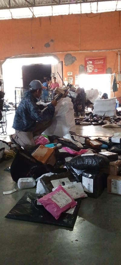 In the city of Kiangroke, Indonesia, inside a warehouse with a corrugated metal roof and an orange-painted wall, a person wearing a camouflage jacket and a baseball cap is kneeling and sorting through a large pile of packages. The packages are of various sizes and shapes, wrapped in plastic and cardboard, and are scattered across the concrete floor. Some packages are black, others are brown or pink. In the background, other people are visible, some of whom appear to be working with the packages as well. A large white plastic bag is positioned behind the kneeling person. A sign with text and QR codes is visible on the orange wall. The scene suggests an active shipping or distribution center.