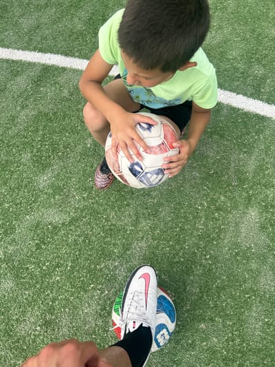 The image shows an overhead view on what appears to be an artificial grass surface, possibly a sports field. In the left portion of the frame, a young boy wearing a light green t-shirt and dark shorts is holding a soccer ball with red, white, and blue panels. The boy's head is angled downward, suggesting he is looking at the ball. Below the soccer ball, the boy is wearing soccer cleats.
To the right, a foot wearing a white Nike soccer shoe with a pink swoosh is resting on top of another soccer ball. A bare arm extends into the frame from the right edge.
A thin white line bisects the top-left portion of the image. The scene is likely outdoors, given the lighting. There is no information to establish a specific connection to the city of Kalchevo, Bulgaria.