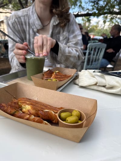 The image depicts an outdoor dining scene within the Emek Izrael Regional Council, Israel. In the midground, an individual, whose torso, arms, and lower face are visible, is seated at a white table. This person wears a light plaid shirt over a white garment with dark long sleeves, and a thin dark necklace. Their hands are actively engaged with a clear plastic cup filled with a green liquid; the right hand presses down on the clear lid, while the left hand holds a dark straw inserted through the lid.
A metal tray positioned on the white table holds the plastic cup and a brown cardboard food tray containing a toasted croissant, sliced and revealing a cheese filling, alongside a small paper cup holding several green olives. In the immediate foreground, another identical brown cardboard food tray rests directly on the white table, featuring a similar toasted croissant and a small paper cup with green olives. The background is an out-of-focus outdoor setting under natural light, revealing tree foliage and a blurred male figure with a beard, dressed in a dark shirt and jeans, seated at a light blue wooden chair.